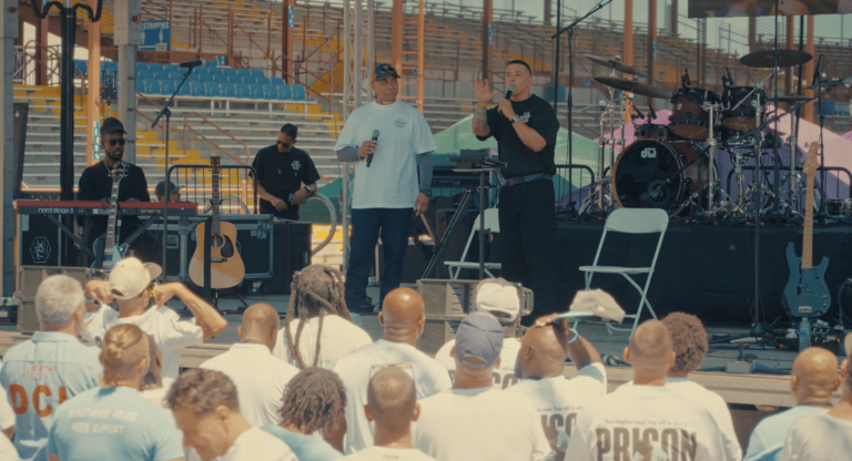 Traduit par un pasteur, Daddy Yankee se tient debout sur une scène et prêche devant des détenus également debout, de dos.