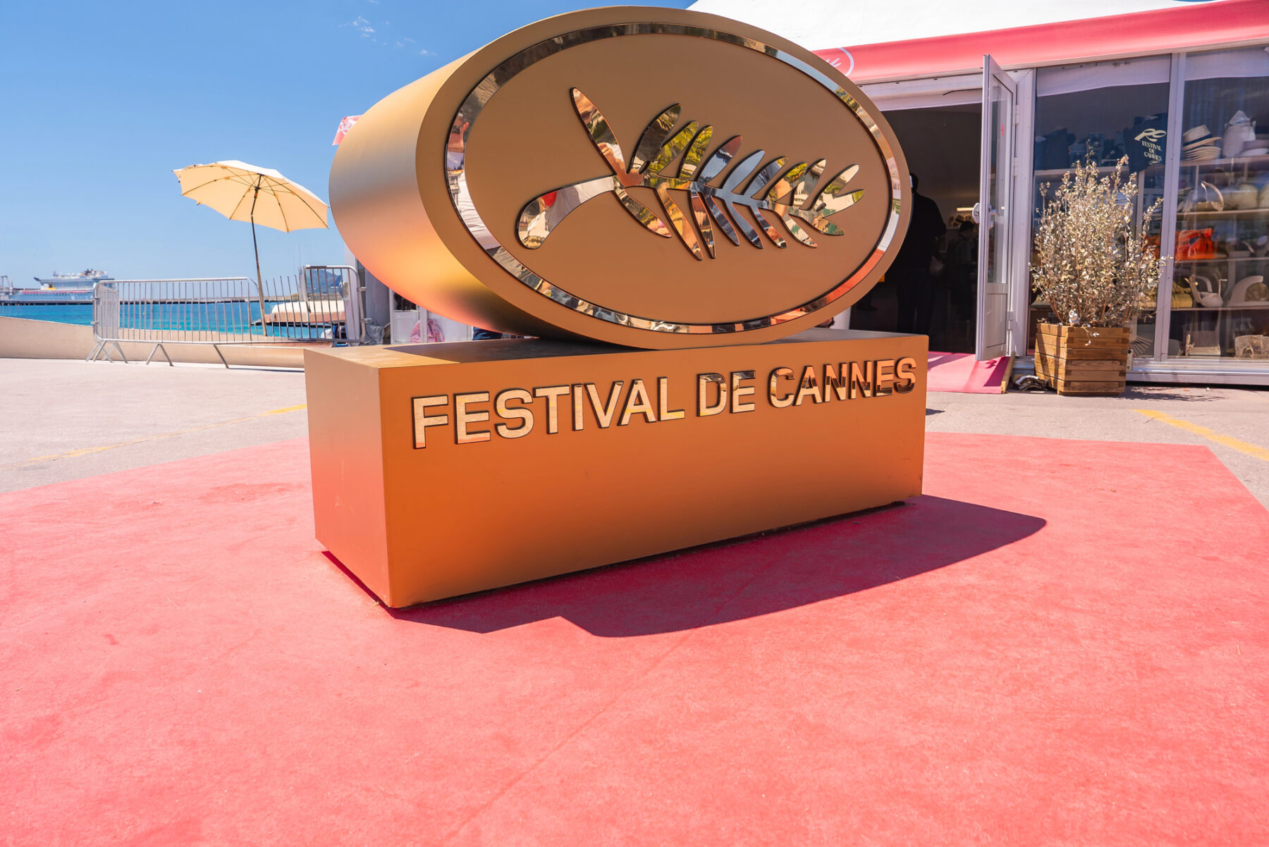 Sculpture de la Palme d'Or dorée sur un tapis rouge, avec l'inscription « Festival de Cannes », installée en extérieur avec un ciel bleu, un parasol, le bord de mer et un immeuble à proximité.