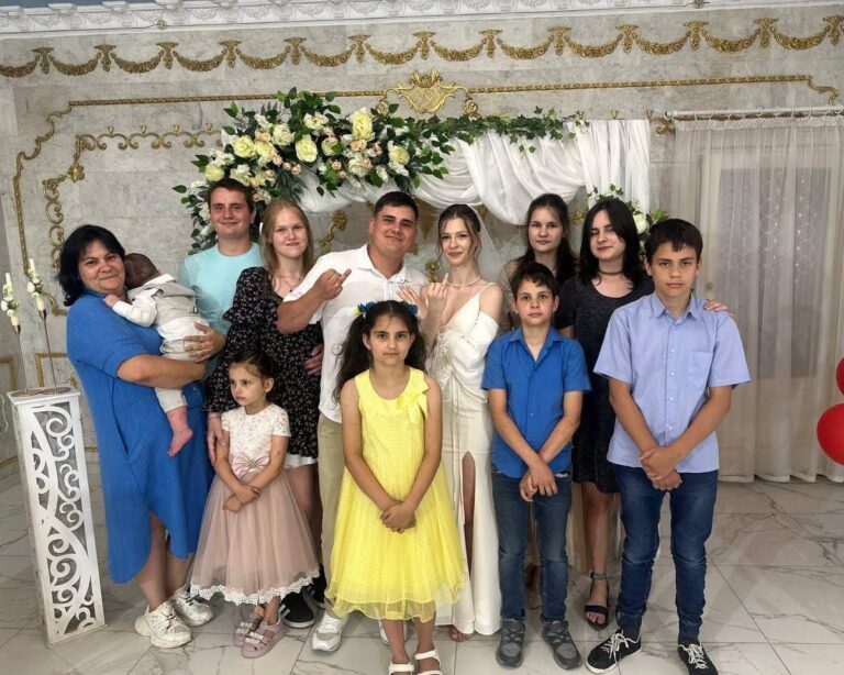 Photographie de la famille Voronov réunie lors d'une célébration de mariage. Onze membres de la famille de différentes générations, incluant des enfants, des adolescents et un bébé, posent ensemble en souriant devant une arche décorée de fleurs blanches. Au centre se tiennent un homme et une femme en tenue de mariés.