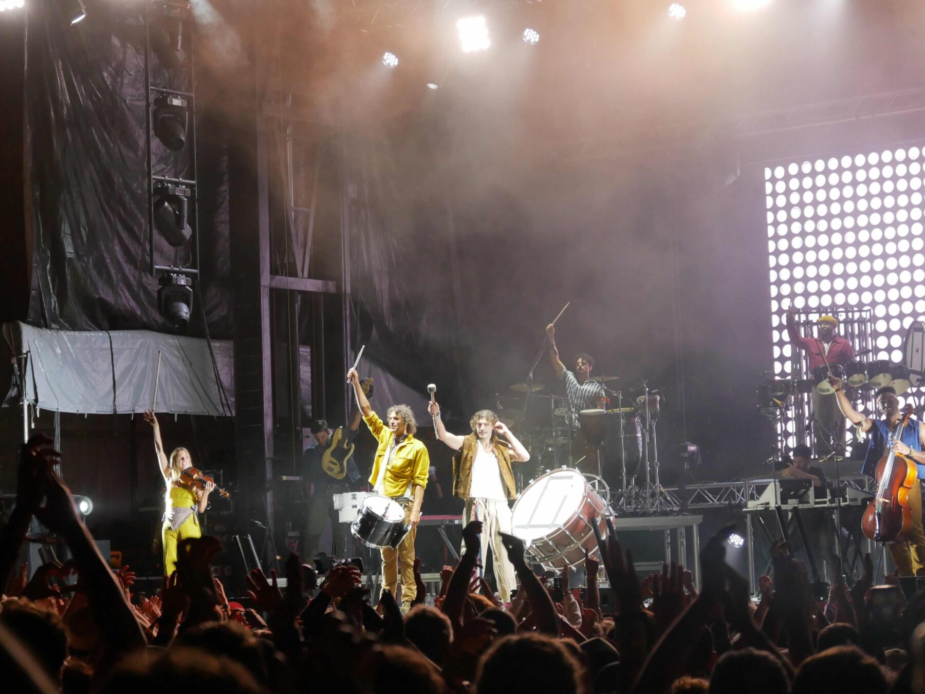 Photographie du groupe de pop-rock chrétien For King & Country en plein concert de nuit sur la scène extérieure du Jesus Festival. Sous les faisceaux lumineux des projecteurs, les deux chanteurs principaux se tiennent au centre de la scène, tenant des baguettes et frappant sur de gros tambours. Ils sont entourés de leurs musiciens, dont une violoniste à gauche et un violoncelliste à droite. Au premier plan, on aperçoit la silhouette d'une foule dense de festivaliers, les bras levés vers la scène dans une ambiance festive.
