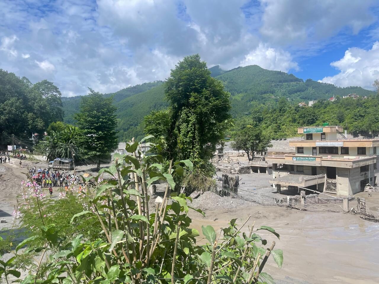 Paysage dévasté par une importante coulée de boue. Au premier plan, une épaisse couche de boue grise et d'eau trouble recouvre la vallée, engloutissant partiellement un bâtiment commercial sur la droite. Sur la gauche, un groupe de personnes est rassemblé pour observer les dégâts au milieu des débris. En arrière-plan, des montagnes verdoyantes sous un ciel bleu nuageux contrastent fortement avec la zone sinistrée.