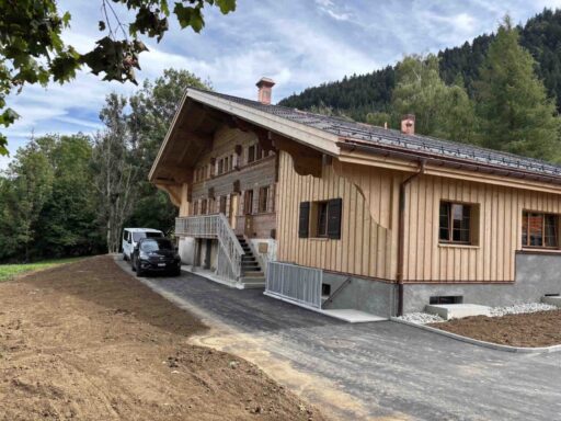 Grand chalet traditionnel suisse récemment rénové, mêlant anciens madriers patinés et extensions en bois clair. Un escalier extérieur mène à une coursive au premier étage, tandis qu'une allée fraîchement goudronnée borde la façade, devant un écrin de forêt de montagne.