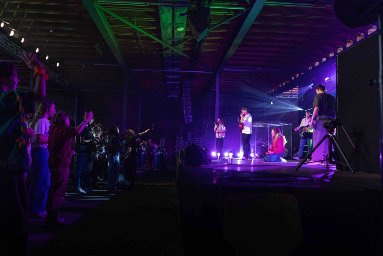 Participants les mains levées face à une scène éclairée en violet, où un groupe de louange joue et une chanteuse prie à genoux, lors de One’ 2025 à l’Espace Gruyère.