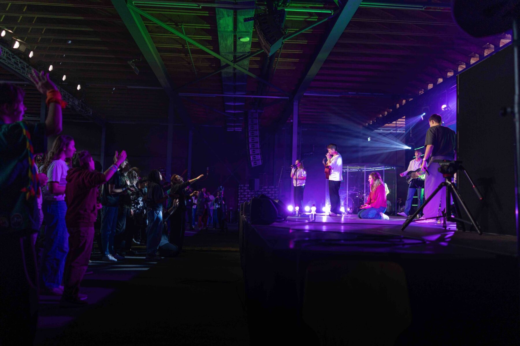 Participants les mains levées face à une scène éclairée en violet, où un groupe de louange joue et une chanteuse prie à genoux, lors de One’ 2025 à l’Espace Gruyère.