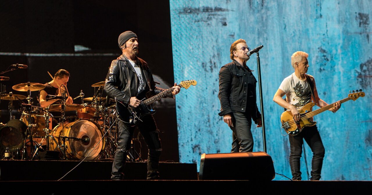 Les quatre membres de U2 sur scène devant un écran bleu: de gauche à droite, Larry Mullen Jr. à la batterie, The Edge à la guitare, Bono au micro et Adam Clayton à la basse