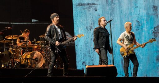 Les quatre membres de U2 sur scène devant un écran bleu: de gauche à droite, Larry Mullen Jr. à la batterie, The Edge à la guitare, Bono au micro et Adam Clayton à la basse