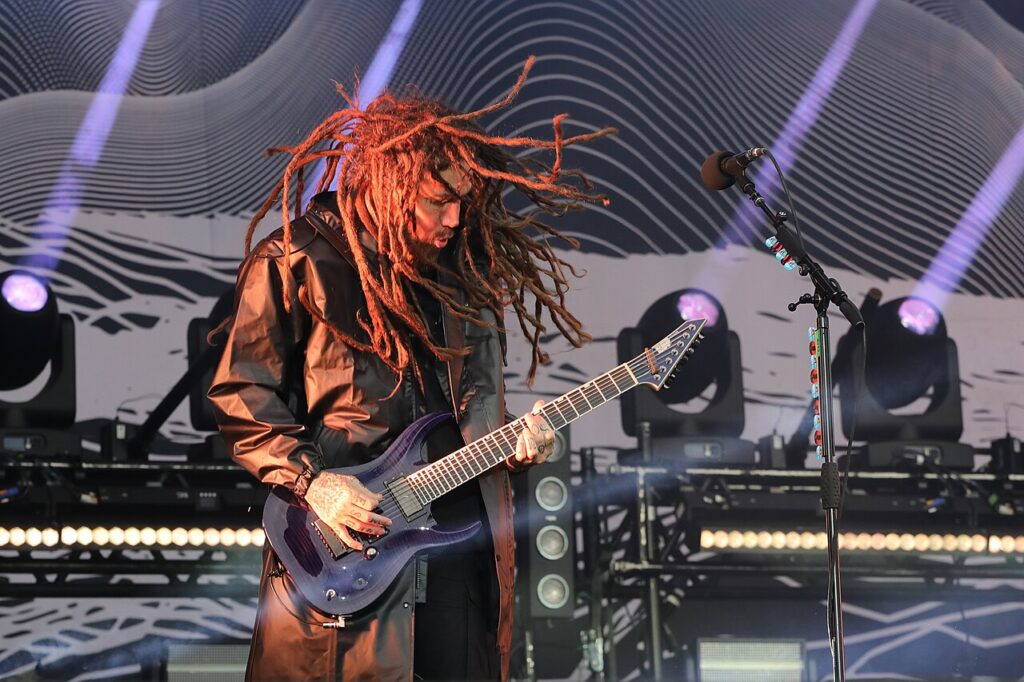 Brian Welch, longues dreadlocks en mouvement et long manteau brun, joue d’une guitare électrique violette sur une scène éclairée par des projecteurs