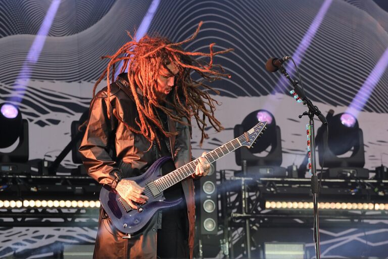 Brian Welch, longues dreadlocks en mouvement et long manteau brun, joue d’une guitare électrique violette sur une scène éclairée par des projecteurs