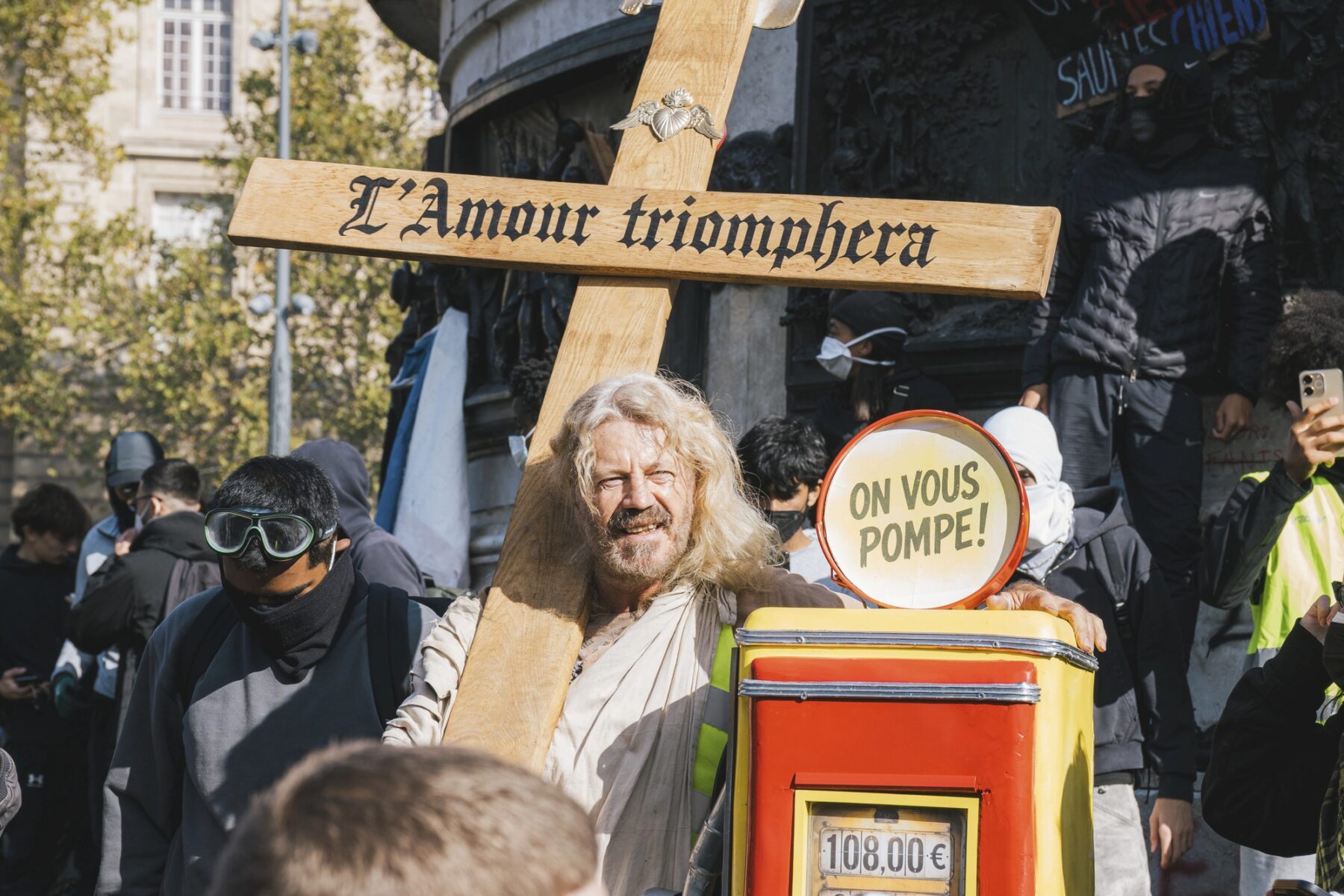Un homme aux cheveux longs et à la barbe porte une grande croix en bois avec l'inscription « L'Amour triomphera » au milieu d'une foule de manifestants. Il tient également une réplique de pompe à essence rouge et jaune sur laquelle est fixé un panneau circulaire indiquant « ON VOUS POMPE ! »